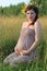 Beautiful pregnant woman in wreath sits in dry field at