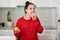 Beautiful pregnant young woman makes choice between fresh apple and doughnut, wears red oversized sweater, poses against kitchen