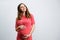 Beautiful pregnant young woman standing over grey wall. Copy space.