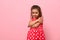 Beautiful preschool 4 years kid girl shyly hugging herself, cute smiling posing on camera against pink wall background with copy