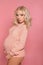 Beautiful pretty pregnant woman expecting maternity hugging her belly. Blonde in knit sweater isolated on studio pink background.