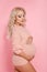 Beautiful pretty pregnant woman expecting maternity hugging her belly. Blonde in knit sweater isolated on studio pink background.