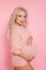 Beautiful pretty pregnant woman expecting maternity hugging her belly. Blonde in knit sweater isolated on studio pink background.