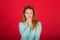 Beautiful red-haired girl closes her mouth with her hand, secret, isolated studio photo on a red background