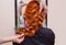 Beautiful, red-haired girl with long hair, hairdresser weaves a French braid, in a beauty salon