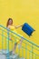 Beautiful red-haired smiling woman joyfully descends the stairs and waving a blue suitcase on a yellow background