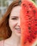 Beautiful red-haired woman smiling with braces on her teeth covers half of her face with a slice of watermelon outdoors