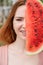 Beautiful red-haired woman smiling with braces on her teeth covers half of her face with a slice of watermelon outdoors