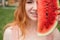 Beautiful red-haired woman smiling with braces on her teeth covers half of her face with a slice of watermelon outdoors