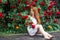 Beautiful redhead girl with bare feet wearing in a white stylish dress sitting on background of blooming roses and holding a