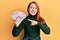 Beautiful redhead woman holding 50 mexican pesos banknotes smiling happy pointing with hand and finger