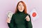 Beautiful redhead woman holding 5000 japanese yen banknotes and japan flag making fish face with mouth and squinting eyes, crazy