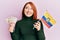Beautiful redhead woman holding colombia flag and 10 colombian pesos banknotes smiling looking to the side and staring away