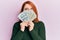Beautiful redhead woman holding dollars close to face clueless and confused expression