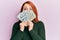 Beautiful redhead woman holding dollars close to face depressed and worry for distress, crying angry and afraid