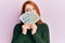 Beautiful redhead woman holding dollars close to face in shock face, looking skeptical and sarcastic, surprised with open mouth