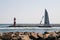 Beautiful sailboat on a smooth water harbor sails past two stone boulder walls going towards a small red and white lighthouse