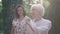 Beautiful senior elegant woman standing in sunrays in spring summer park with blurred granddaughter touching shoulder in