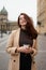 Beautiful serious stylish fashionable smart girl holding cup of coffee in hands and smiles, goes walking down street of St.