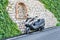 A beautiful shiny black motorbike with reflections is on the side of the road by an old gray stone wall with an orange arch and