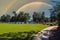 A beautiful shot of a park on the lake with vast green grass, blue lake water, lush green trees and blue sky with a rainbow