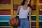 beautiful small young girl in white tank top and denim blue jeans retro fashion holding basketball ball on color wooden