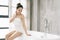 Beautiful smiling Asian woman is relaxing sitting at the side of freestanding white tub in bathroom. Favorite bathing rituals,