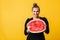 Beautiful smiling girl in black dress happily looking in camera while holding half of watermelon in hands over yellow