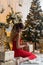 Beautiful smiling girl in evening attire near the New Year tree and with gifts. Festive hairstyle and makeup