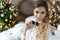 Beautiful smiling girl in evening attire near the New Year tree and with gifts. Festive hairstyle and makeup