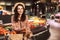 Beautiful smiling girl in hat leaning on trolley full of products happily looking in camera with cellphone in hand in