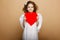 Beautiful smiling little curly girl with wings, in dress, holding a red heart, looking at camera, over beige background.