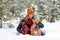 Beautiful smiling woman is sitting on a snow with her two dogs wrapped in red checkered plaid in winter coniferous forest