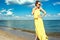 Beautiful smiling woman with updo hair wearing long yellow striped baggy summer dress and round sunglasses walking along the sea