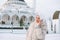 Beautiful smiling young Muslim woman in headscarf in light clothing using mobile against the background of mosque in the winter