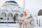 Beautiful smiling young Muslim woman in headscarf in light clothing using mobile against the background of mosque in the winter