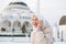 Beautiful smiling young Muslim woman in headscarf in light clothing using mobile against the background of mosque in the winter