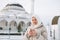 Beautiful smiling young Muslim woman in headscarf in light clothing using mobile against the background of mosque in the winter