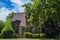 Beautiful stone two story cottage with green shutters and a steep roof surrounded by lush vegetation and shadowed by a huge tree