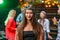 Beautiful stylish caucasian brunette woman in a bandanna looks at the camera while her friends dancing in colored disco