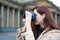 Beautiful stylish fashionable girl holds camera in her hands and takes pictures. Woman photographer with long dark hair in city,