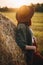 Beautiful stylish woman in hat looking at sunset light at haystacks in summer field.Tranquility