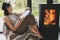 Beautiful stylish woman reading book on chair at warm burning fireplace, cozy vacation. Leisure time