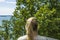 Beautiful summer Baltic sea landscape view. Young blond woman looking on sea through green tree tops