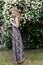 Beautiful tender blonde girl in a long dress with evening hairstyle standing in the garden near a flowering aromatic tree