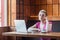 Beautiful thinkful young girl freelancer with short hair, in pink t-shirt and eyeglasses is sitting in cafe and working on laptop