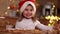 Beautiful three year girl in santa hat sitting by the table in front of kitchen decorated fairy lights