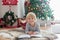 Beautiful toddler child, reading book front of  Christmas tree, decoration and presents around him