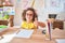 Beautiful toddler wearing glasses and unicorn diadem sitting on desk at kindergarten doing happy thumbs up gesture with hand