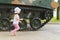 A beautiful two-year-old girl stands on a Sunny day on the street near the huge wheels of a tank, side view. A child next to a tan